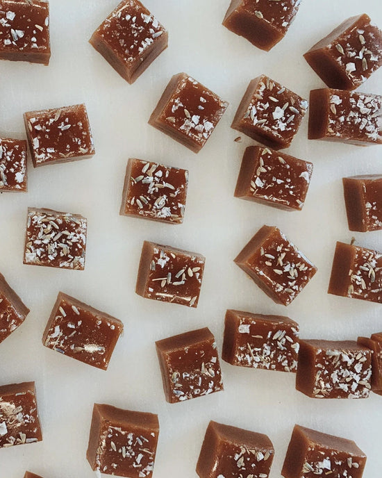 Several square Honey Lavender Caramels by Wildflower Caramel Co are arranged in a scattered pattern on a white surface, with some sprinkled with flaky sea salt.
