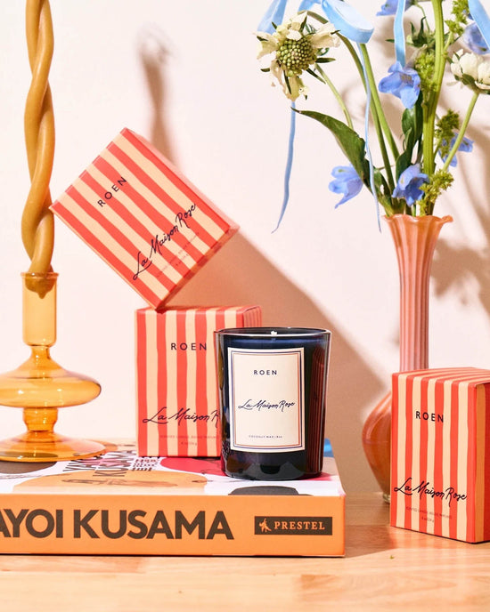 A black ROEN La Maison Rose Candle made from coconut soy wax sits on a Yayoi Kusama book, surrounded by red-striped boxes, a gold candle holder, and a vase with blue and white flowers.