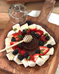 A wooden platter with brie cheese wedges, strawberries, blackberries, and basil leaves surrounds an etúHOME Bistro Glass Jam Jar, Round filled with honey and a honey dipper.