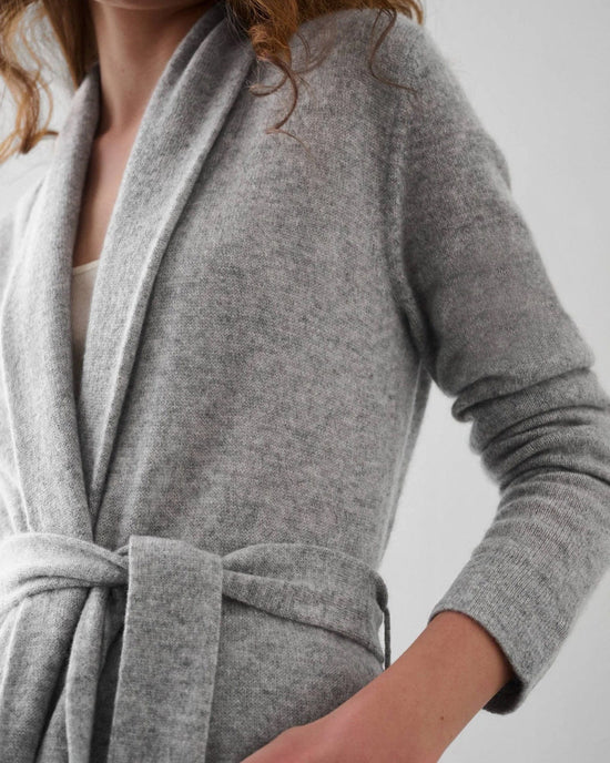 A person wearing the White + Warren Cashmere Long Robe in Grey Heather, belted with long sleeves and an oversized fit over a white top, wavy brown hair partially visible, stands against a plain light background.