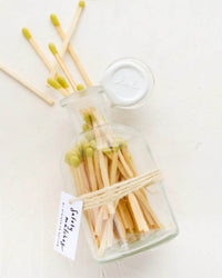 The LEIF Apothecary Jar Matches w/ Wax Seal in Citron features green-tipped matches in a reusable glass jar, sealed with a white cap, twine, and “Safety Matches” tag. Some matches are scattered beside the vibrant jar.