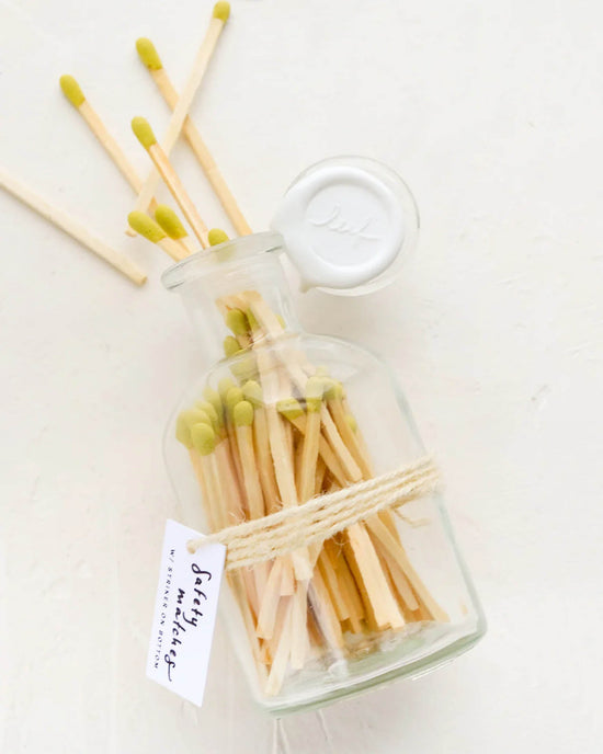 The LEIF Apothecary Jar Matches w/ Wax Seal in Citron features green-tipped matches in a reusable glass jar, sealed with a white cap, twine, and “Safety Matches” tag. Some matches are scattered beside the vibrant jar.