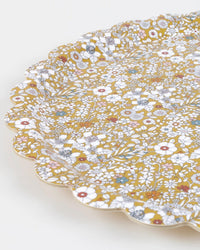 The Meri Meri x Liberty June's Meadow Small Tray features a scalloped edge and a dense floral pattern in white, yellow, pink, blue, and green on a mustard background, covering the entire surface.