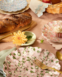 A table set with Meri Meri X Liberty Mixed Set Reusable Large Plates by Meri Meri, a gold fork, bread on a wooden board, a yellow flower, floral cupcake, napkins, fruit basket, all atop a pink tablecloth.