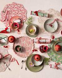 A festive table set with Meri Meri x Liberty Winter Placemats S/4, pomegranates, pink and green ribbon-tied crackers, assorted cutlery and glasses on a white cloth, adorned with small flowers and berries.