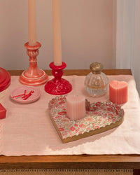A wooden table with a pink cloth holds two Meri Meri X Liberty Scalloped Heart Candles in holders, a round glass perfume bottle, a tray with a floral heart pattern—ideal for Valentine’s—a pink ribbed candle, matchsticks in a dish, and a pink square candle.