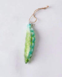 The Cody Foster Garden Pea - Mid Green Ornament is a hand-crafted glass decoration shaped like a green pea pod with sparkling accents, hanging from brown twine against a light gray background.