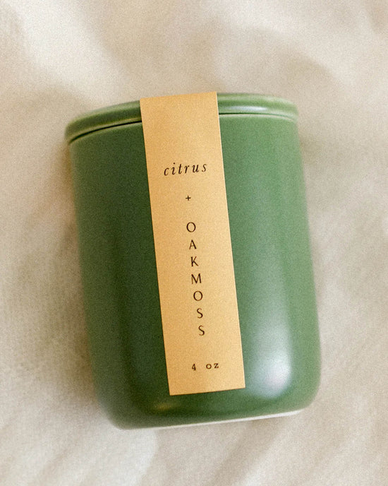 The LEIF Citrus & Oakmoss Candle, made from a soy wax blend and weighing 4 oz, comes in a green jar with a beige label and is shown resting on a light-colored textured surface.