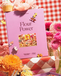 The Piecework Puzzles Flour Power - Better Homes & Gardens Collab 1000 Piece Puzzle exudes home-baked charm, with colorful flowers, vintage cookbooks, pink drinks, a red checkered tablecloth, and scattered puzzle pieces surrounding the box.