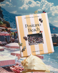 Displayed outdoors on a large seashell, the Piecework Puzzles Positano Beach - Slim Aarons Collab 1000 Piece Puzzle is shown with loose pieces, set against a scenic Amalfi Coast beach and rocky coastline background.