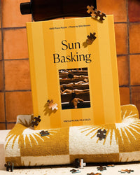 A Piecework Puzzles Sun Basking - Slim Aarons Collab 1000 Piece Puzzle box stands upright on a rolled yellow-and-white towel, with loose pieces scattered around, set against brown tiled walls reminiscent of Slim Aarons photography.