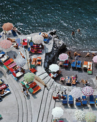 Aerial view of sunbathers on colorful loungers and umbrellas by the dark blue sea, featured in the "Positano Beach - Slim Aarons Collab 1000 Piece Puzzle" by Piecework Puzzles. Some relax on stone steps, evoking a classic Slim Aarons scene.