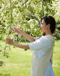 A woman wearing The Floral Society's Canvas Workshop Apron in Natural stands outdoors, gently touching white flowering tree branches amid sunlight and greenery.