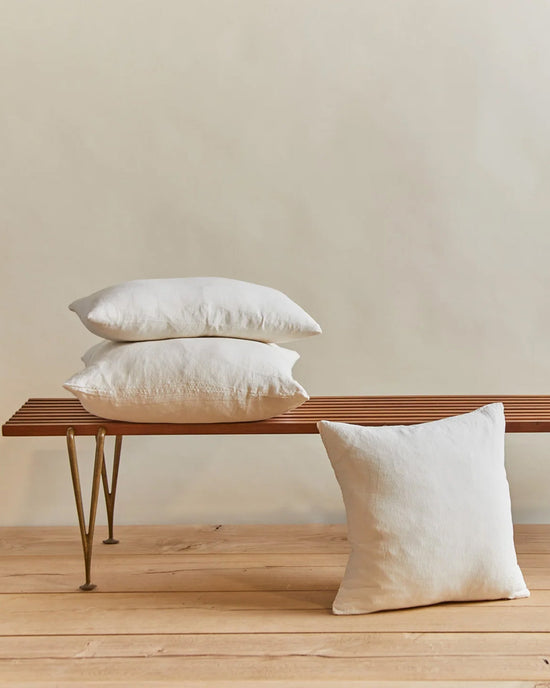 Three Hawkins New York Simple Linen Pillows (22x12 Ivory) are stacked on a slatted wooden bench with metal legs, while another rests on the light wood floor. Each features a soft down insert and is machine washable for easy care.