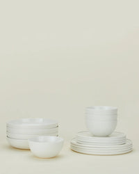 The Hawkins New York Simple Porcelain Dinnerware 16 Piece Set features neatly stacked plates and bowls, all in classic white, elegantly arranged against a light beige background.