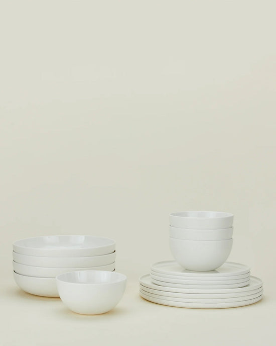 The Hawkins New York Simple Porcelain Dinnerware 16 Piece Set features neatly stacked plates and bowls, all in classic white, elegantly arranged against a light beige background.