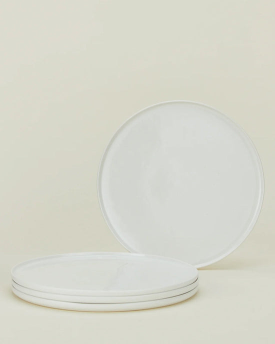 Four Simple Porcelain Dinner Plates by Hawkins New York are displayed on a light beige background—three stacked horizontally and one propped vertically behind—highlighting their elegant fine porcelain style.