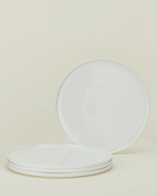 Four Simple Porcelain Dinnerware plates from Hawkins New York are displayed on a neutral background; three are stacked and one stands upright behind, highlighting this minimalist 16-piece set.