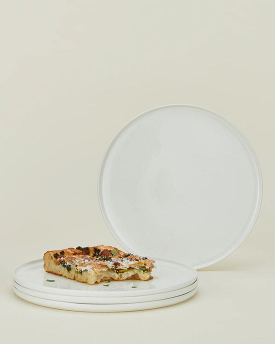 A stack of three Simple Porcelain Dinner Plates from Hawkins New York holds a rectangular slice of pizza, while a fourth plate from the set of 4 is propped upright behind them against a light beige background.