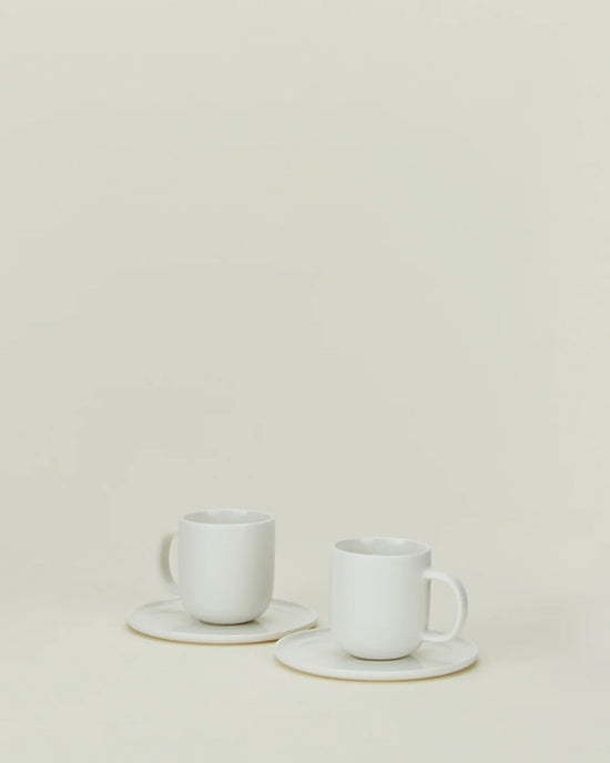 Two Simple Porcelain Espresso Cups & Saucers by Hawkins New York rest on a neutral background, their minimalist design and fine porcelain elegance enhanced by the surrounding empty space. Set includes 2 cups and saucers.