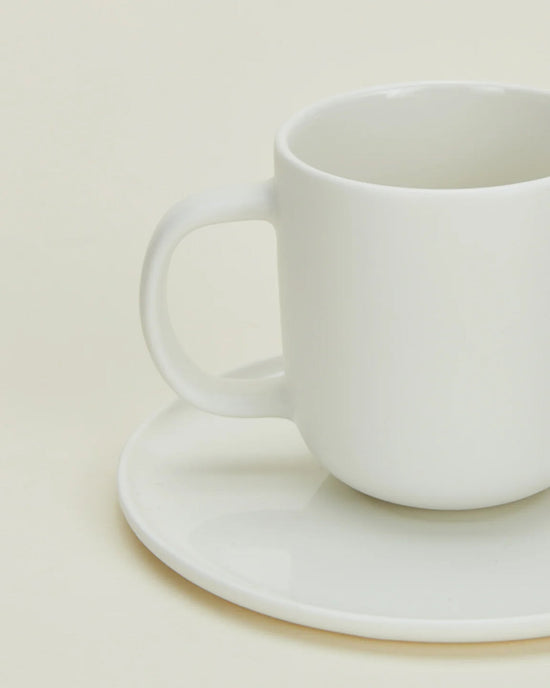A Simple Porcelain Espresso Cup & Saucer from Hawkins New York sits empty and slightly off-center, its handle on the left, against a light cream background—showcasing minimalist elegance from this stylish set of 2.