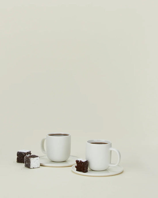 The Simple Porcelain Espresso Cups & Saucers - Set of 2 by Hawkins New York are shown on a plain light background, each on its saucer and paired with a chocolate brownie and a sugar-dusted brownie.