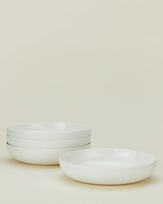Four Simple Porcelain Low Bowls - Set of 4 from Hawkins New York are stacked on the left, with one more bowl in front right, all set against a light cream background—ideal for displaying elegant porcelain dinnerware.