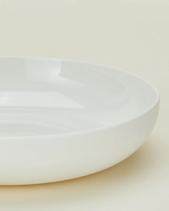A close-up of a Simple Porcelain Low Bowl from Hawkins New York, part of a set of 4, shown from the side against a neutral background, highlighting its smooth surface and gently raised rim.