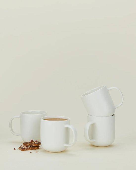 Four Hawkins New York Simple Porcelain Mugs—set of 4—are shown on a light background; two are stacked, one holds coffee, and one is empty with chocolate pieces scattered nearby.