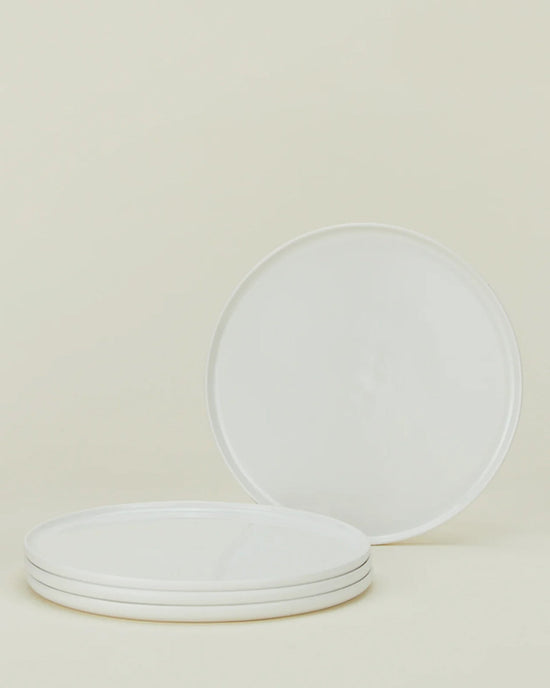 Four round, white plates from the Hawkins New York Simple Porcelain Dinnerware 16 Piece Set are arranged on a neutral background—three stacked and one upright to show its smooth, plain surface.
