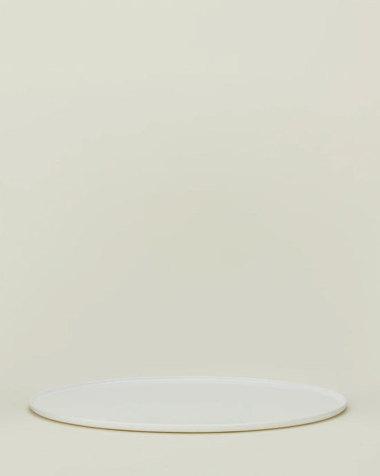 The Simple Porcelain Serving Platter by Hawkins New York, a round white porcelain piece, rests on a light beige background with empty space above, reflecting the brand’s minimalist elegance.