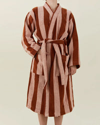 A person wears the Hawkins New York Simple Striped Terry Bathrobe in Blush/Terracotta, a knee-length, long-sleeved robe with wide vertical brown and pink stripes and a matching waist belt. The person’s face is not visible.