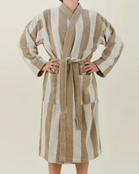 A person in the Hawkins New York Simple Striped Terry Bathrobe in Ivory/Flax, made from 100% cotton, stands with hands on hips against a plain light background.