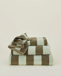 A stack of three Simple Striped Terry Hand Towels in Olive/Sage by Hawkins New York is shown against a plain, light background, with the smallest towel draped over the top. Made in Portugal.