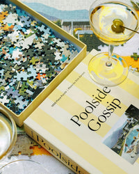 A partially completed Piecework Puzzles Poolside Gossip – Slim Aarons Collab 1000 Piece Puzzle sits beside its box, with two glasses of white wine and green olives on the table among scattered pieces for a midcentury vibe.