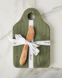 The etúHOME Dutch Small Cheese Board w/ Spreader in Spruce Green features a textured green finish and includes a wooden spreader, both tied with a white ribbon and displayed on a marble surface.