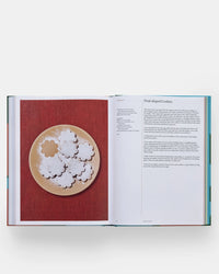 An open copy of The Spanish Mediterranean Islands Cookbook by Phaidon Press displays petal-shaped cookies; the left page shows Mediterranean cookies dusted with powdered sugar, while the right page features the recipe.