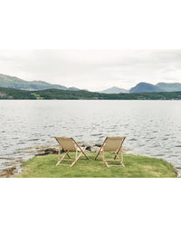Two empty deck chairs labeled "Outdoor Chic" by Cozy Publishing sit on grass at the edge of a calm lake, facing distant green hills and mountains beneath a cloudy sky.